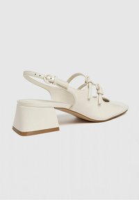 Chaussure slingback à bout pointu de couleur crème, ornée de deux nœuds décoratifs sur le dessus et d'un talon bloc avec un contour de semelle marron.