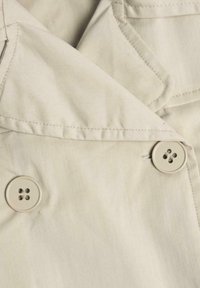 Světle béžová trench coat látka s dvouduchým designem, dvěma velkými knoflíky a klopovým límcem, hladkou texturou a minimálním zdobením.