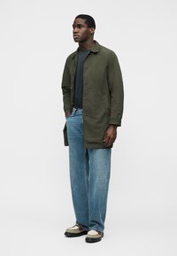 Muž stojaci, oblečený v olivovo zelenom kabáte na gombíky, čiernom tričku, voľných modrých džínsoch a béžovo-hnedých topánkach, na jednoduchom svetlom pozadí.