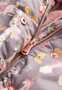 Gros plan d'une fermeture éclair en or rose rose et d'un bouton-pression sur une veste grise avec des motifs colorés de papillon, lune, étoile, arc-en-ciel et licorne.