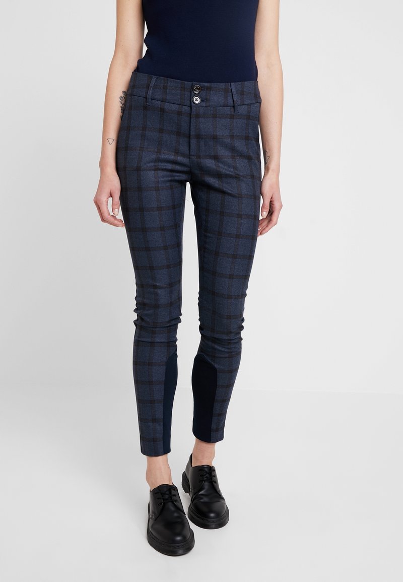 Femme portant un pantalon slim bleu à carreaux et des chaussures noires à lacets, debout devant un fond blanc uni.