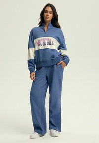 Niebieski zestaw sportowy składający się z bluzy z pół-zamkiem z napisem "MICHIGAN" w różowym kolorze, w połączeniu z szerokimi niebieskimi spodniami. Miękka tkanina z luźnym krojem.