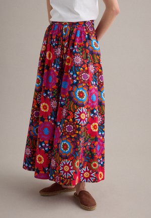 Persona que lleva una falda larga y colorida con estampado floral combinada con una parte superior blanca y alpargatas marrones sin cordones sobre un fondo neutro.