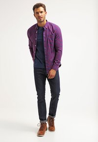 Button-up checkered shirt in purple and blue over a navy t-shirt, paired with dark jeans and brown lace-up boots.