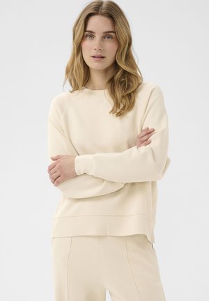 Femme aux longs cheveux blonds ondulés portant un sweat-shirt couleur crème et un pantalon assorti, debout les bras croisés devant un fond uni.