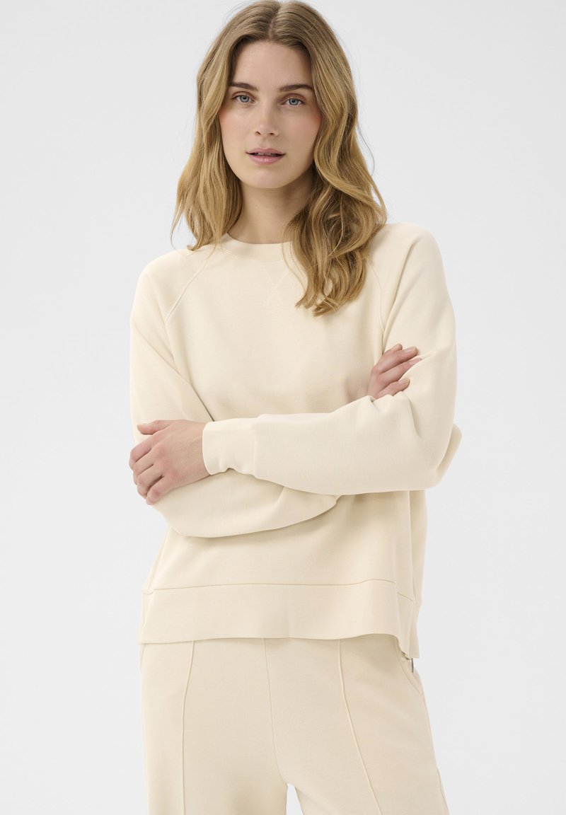Femme aux longs cheveux blonds ondulés portant un sweat-shirt couleur crème et un pantalon assorti, debout les bras croisés devant un fond uni.