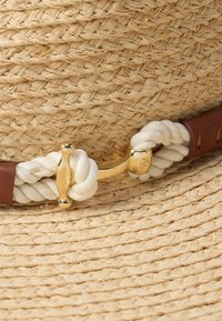 Καπέλο από ψάθα με πλεγμένο σχέδιο, μπεζ χρώμα, διακοσμημένο με λευκή ταινία σχοινιού και χρυσά μεταλλικά στοιχεία. Καφέ δερμάτινο λουρί στερεώνει την ταινία.