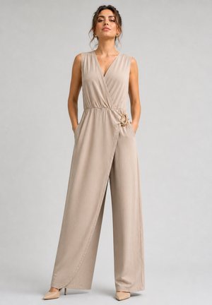 Femme portant une combinaison beige sans manches portefeuille avec jambes larges et un détail anneau sur le côté, debout les mains dans les poches sur un fond uni.