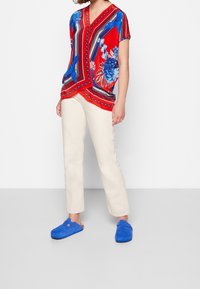 Blouse à motifs rouges avec des accents floraux bleus, associée à un pantalon droit de couleur crème et des sabots en daim bleus.