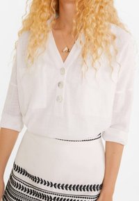 Mujer con pelo rubio rizado que lleva una blusa blanca de botones y una falda blanca con rayas horizontales de un patrón de hojas negras.