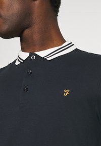 Navy polo shirt with a contrasting white and black striped collar, three buttons, and an embroidered yellow logo on the chest.