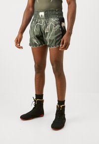 Atlet iført mørkegrønne bokseshorts med hvidt slangemønster og sorte høje bokssko med røde såler, stående på hvidt gulv.