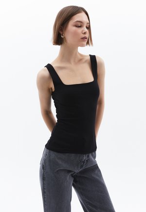 Schwarzes, tailliertes Tanktop mit quadratischem Ausschnitt und breiten Trägern, aus glattem Stoff. Kombiniert mit lockeren grauen Jeans. Minimalistisches Design.