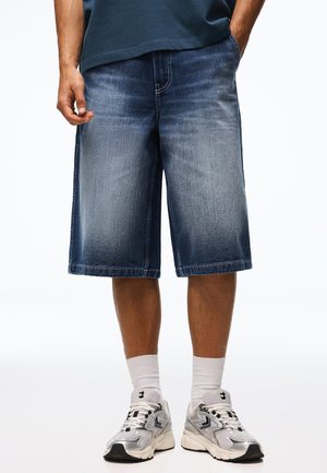 Mann trägt knielange blaue Jeansshorts, weiße Sportsocken und silber-weiße Sportschuhe und steht mit den Händen in den Taschen.