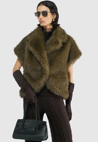 Olijfgroen nepbont cape met korte mouwen, over een donkere gebreide top en bruine kabelbrei broek. Zwarte handtas en bruine handschoenen.