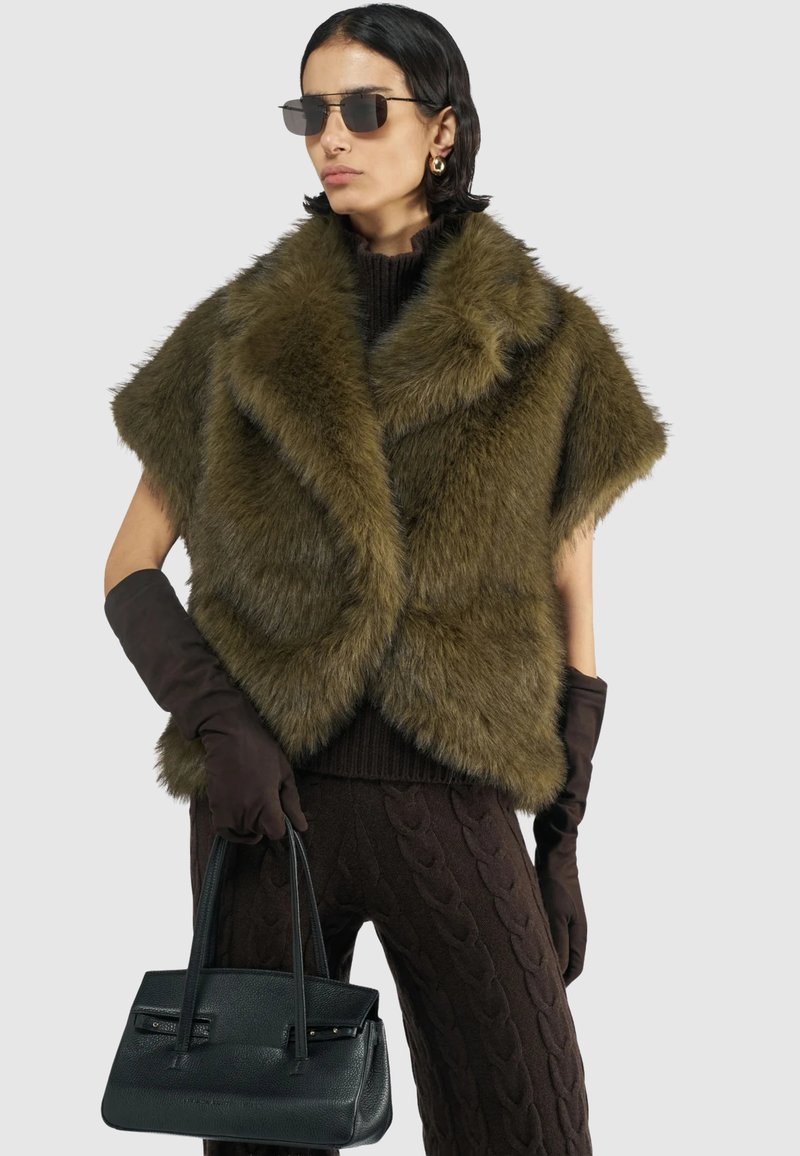 Olijfgroen nepbont cape met korte mouwen, over een donkere gebreide top en bruine kabelbrei broek. Zwarte handtas en bruine handschoenen.