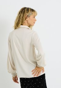 Blusa beige claro con textura suave, que cuenta con cuello y mangas largas abullonadas con puños recogidos. Se lleva con una falda negra texturizada.