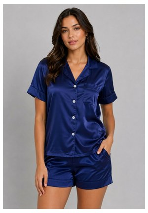 Femme aux longs cheveux foncés porte un haut bleu en satin à manches courtes boutonné et un short assorti, debout avec une main dans la poche devant un fond uni.