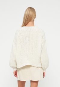 Femme blonde aux cheveux raides portant un pull en maille blanc oversize et une jupe en velours côtelé crème, debout de dos face à un fond blanc.