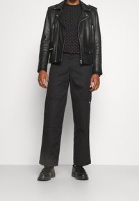 Black leather biker jacket over a dark patterned sweater, paired with wide black trousers and black shoes with a thick sole.