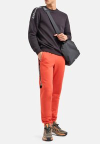 Felpa nera con logo sulla manica, pantaloni della tuta arancioni con striscia laterale e borsa nera; abbinati a scarpe da ginnastica grigie e arancioni.