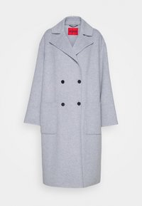Manteau en laine gris clair à double boutonnage avec larges revers, deux boutons noirs, grandes poches frontales et étiquette Hugo rouge à l'intérieur du col.