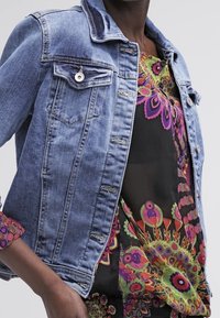 Veste en denim bleu clair avec un col classique, des boutons et deux poches poitrine, portée sur un chemisier coloré à motifs.