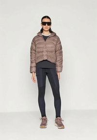 Casaco puffer castanho cropped com fecho frontal e capuz, combinado com leggings pretas com painéis contrastantes e sapatos desportivos castanhos.