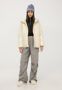 Kurtka puchowa w błyszczącej kremowej kolorystyce, zapinana na zamek, z kapturem. Czarny krótki sweter i szare spodnie z szerokimi nogawkami. Szara czapka beanie z napisem "CREATIVE".