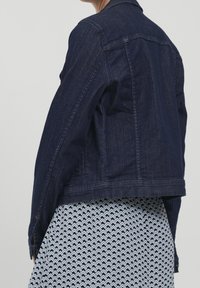 Mörkblå denimjacka med figursydd passform, knappade ärmslut och klassisk krage. Bärs över en mönstrad kjol med geometrisk design.