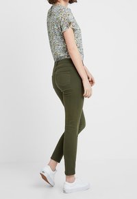 Grüne Skinny-Jeans mit hoher Taille, ausgestattet mit Gesäßtaschen, kombiniert mit einer kurzärmligen blumigen Bluse und weißen Sneakers.
