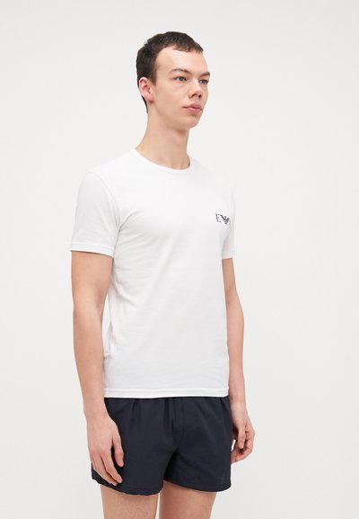 T-shirt en coton blanc à manches courtes, col rond et petit logo noir sur la poitrine gauche, associé à un short foncé.