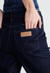 Tamno plavi traperice s kožnom oznakom "Wrangler" i prepoznatljivim šavovima na stražnjim džepovima. Materijal ima glatku teksturu.