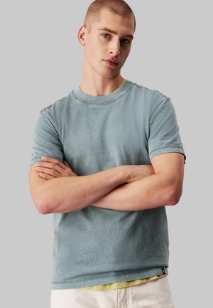 Kortærmet t-shirt med rund hals i dæmpet blå bomuld med synlige stikninger. Lagd over en gul undertrøje, båret med armene korslagt.