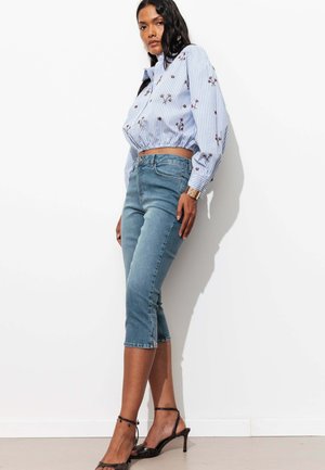 Femme debout contre un mur blanc portant un chemisier court rayé bleu avec broderie florale, un jean capri bleu ajusté et des talons noirs à bride à la cheville.