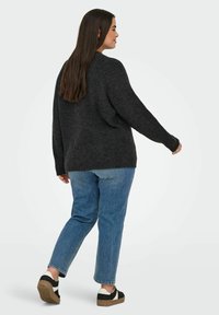 Frau mit langen, dunklen Haaren, die in einem dunkelgrauen Strickpullover, blauen Jeans und schwarz-weißen Sneakers weggeht.