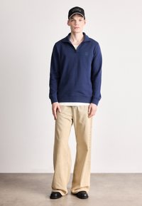 Μπλε σκούρο φούτερ με φερμουάρ και μικρό λογότυπο, σε συνδυασμό με ανοιχτό μπεζ παντελόνι φαρδιάς γραμμής και μαύρα παπούτσια, στέκεται σε ουδέτερο δάπεδο.