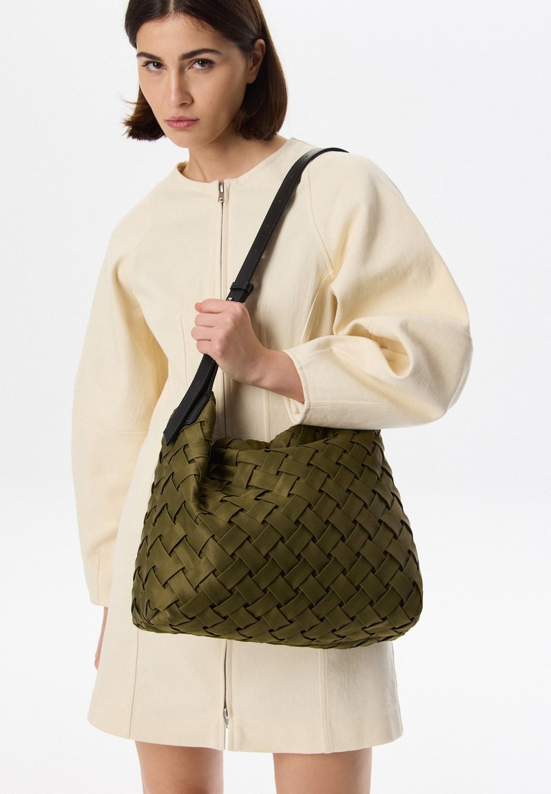 Borsa a tracolla in tessuto verde oliva con una texture morbida, forma curva e tracolla nera regolabile, tenuta da una modella in abito color crema.