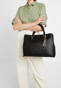 Borsa tote nera realizzata in materiale sintetico testurizzato, con dettagli in metallo dorato e un charm. Forma semplice e strutturata con doppi manici.