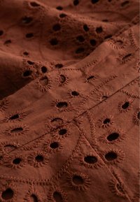 Καφέ ύφασμα με κέντημα eyelet που παρουσιάζει κυκλικές τρύπες και περίπλοκα κεντημένα σχέδια σε μια μαλακή, υφή επιφάνεια.