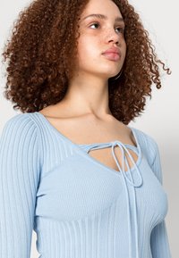 Top de malha canelada azul claro com decote em forma de U e detalhe de laço. Mangas longas e design texturizado realçam o aspeto e o ajuste geral.