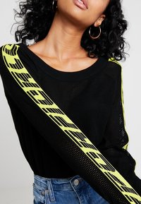Maglione in rete nero con una prominente striscia diagonale gialla neon e accenti grafici neri, abbinato a pantaloni shorts in denim azzurro chiaro.
