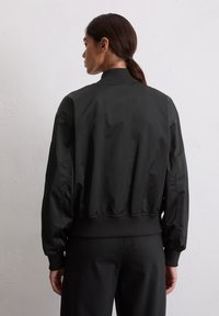 Woman with dark hair in a low ponytail wearing a black bomber jacket and black pants, facing a plain white textured wall.