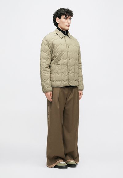 Κουβερτωμένη ανοιχτό μπεζ ζακέτα με λαιμόκοψη για κολάρο, που διαθέτει σχέδια ζιγκ-ζαγκ και μπροστινή τσέπη, σε συνδυασμό με φαρδιά καφέ παντελόνια και πράσινα παπούτσια.