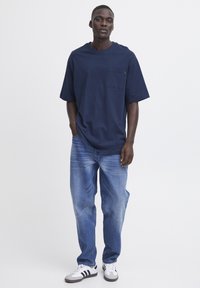 Tricou oversize de culoare bleu marin cu mâneci scurte și un buzunar pe piept, asortat cu blugi de culoare bleu deschis și adidași albi cu dungi negre.