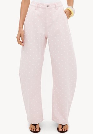 Femme portant un pantalon large rose clair à pois blancs, une main dans la poche, associée à des sandales noires et des bracelets.