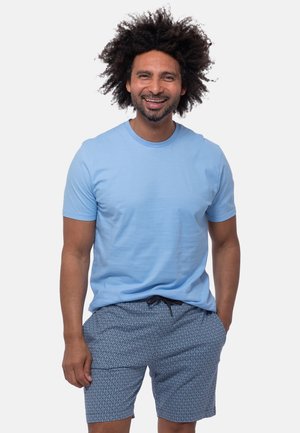 Man with curly hair wearing a light blue t-shirt and patterned blue shorts smiling with one hand in his pocket.