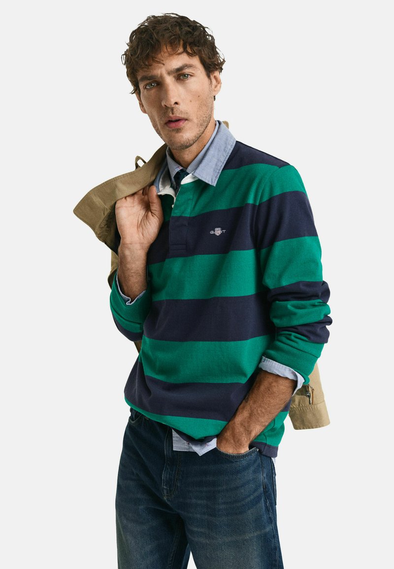 Groen en marineblauw gestreepte lange mouwen polo met een kraaghals, gecombineerd met een lichtblauw overhemd eronder en spijkerbroek.