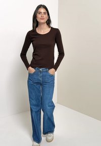 Femme debout contre un mur bicolore, portant un haut marron foncé à manches longues, un jean bleu à jambes larges, des baskets blanches, les mains dans les poches.