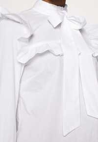 Blusa bianca con dettagli arricciati e colletto a fiocco. Texture liscia con una finitura leggermente lucida. Chiusura con bottoni sul retro.